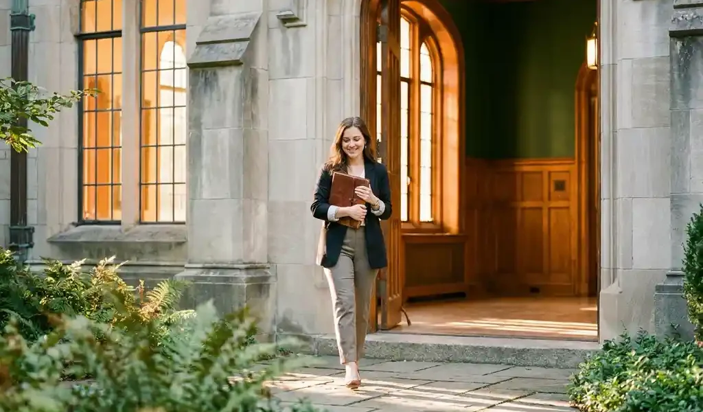 Zelfverzekerde vrouw met leren dagboek tegen haar borst, halverwege een stap op een stenen pad bij een verlichte gebouwsingang tijdens het gouden uur.